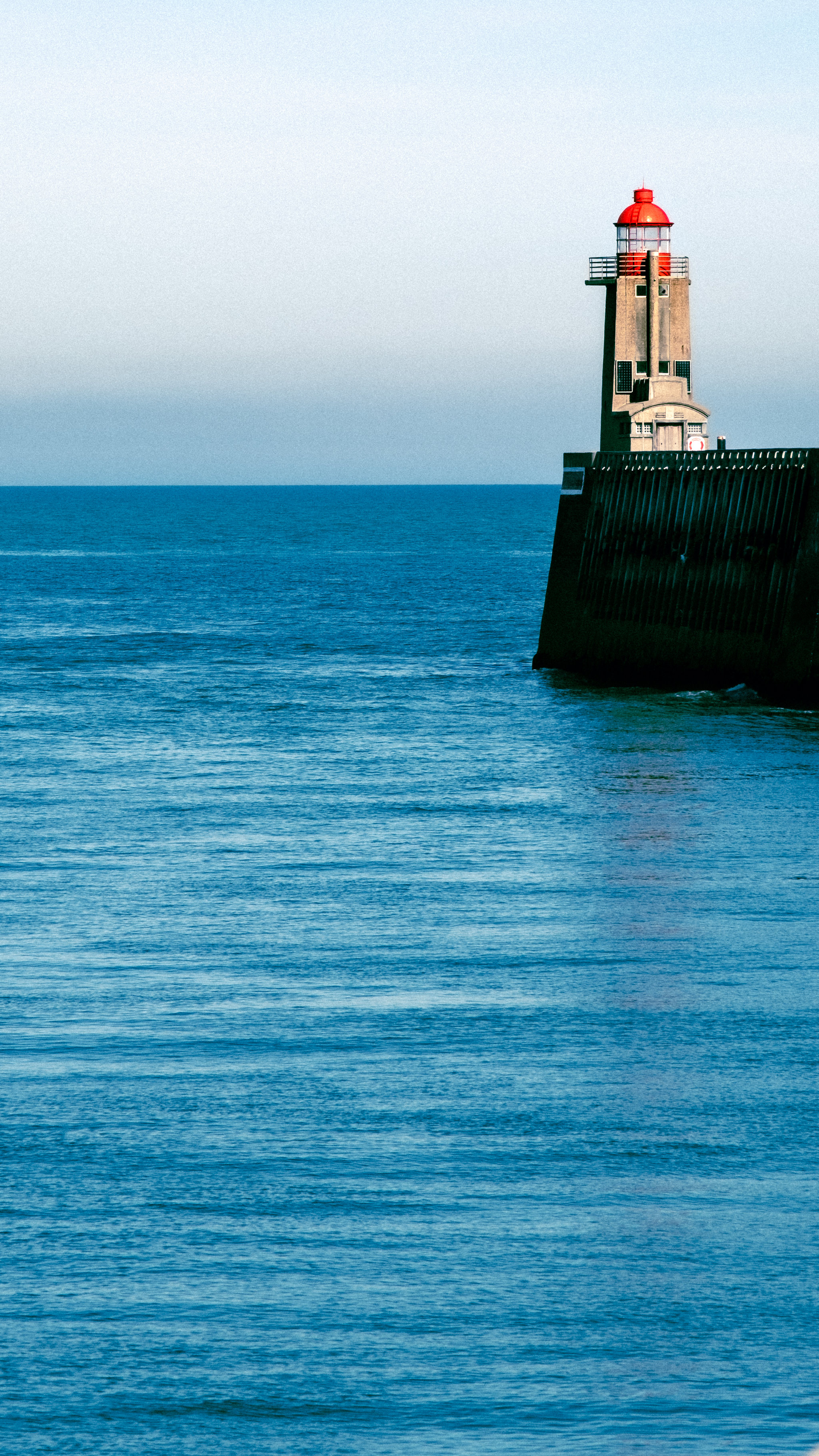 A white lighthouse with a red top rising from a blue sea wall.