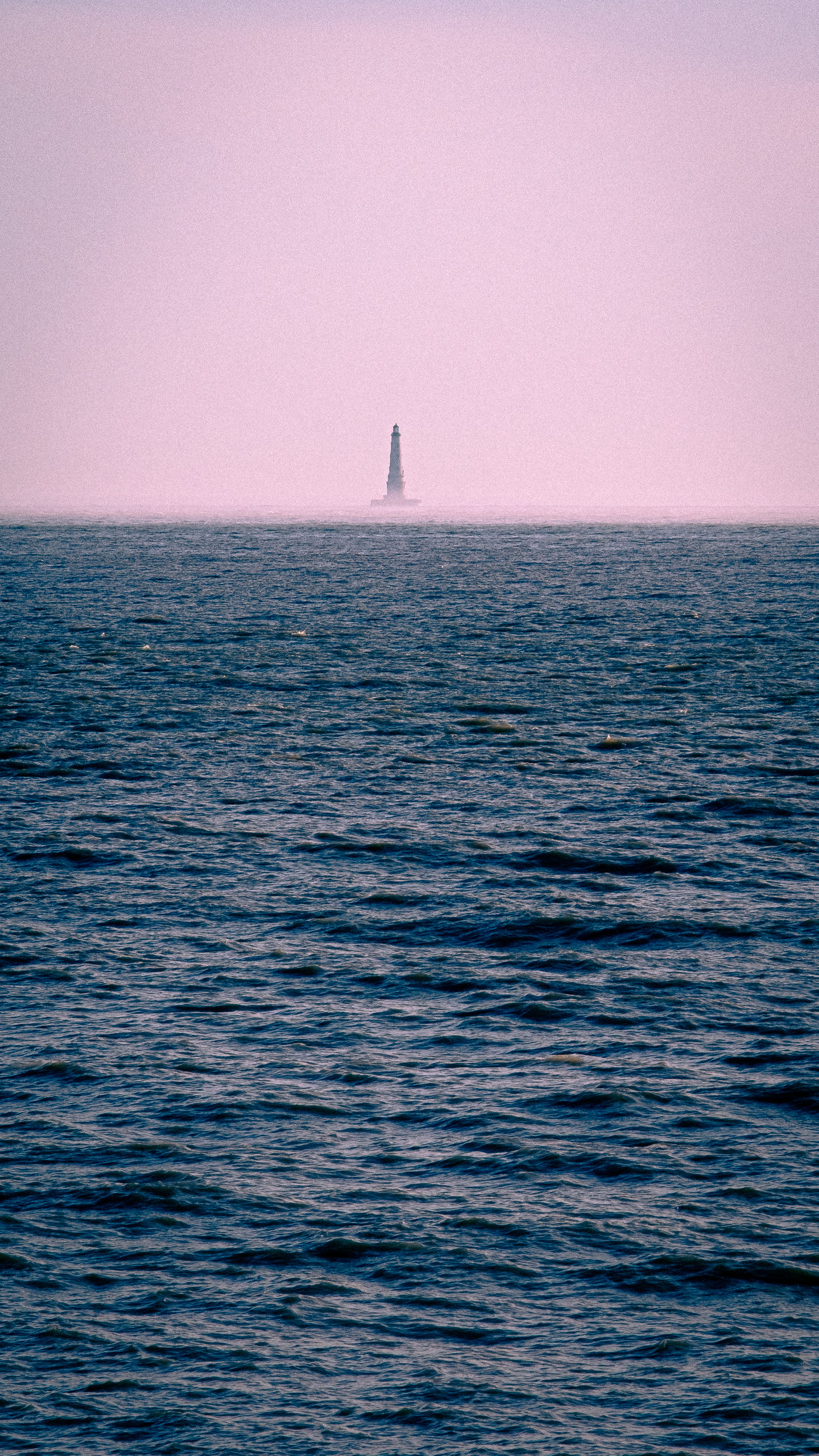 A distant lighthouse on a pale horizon above dark blue water.