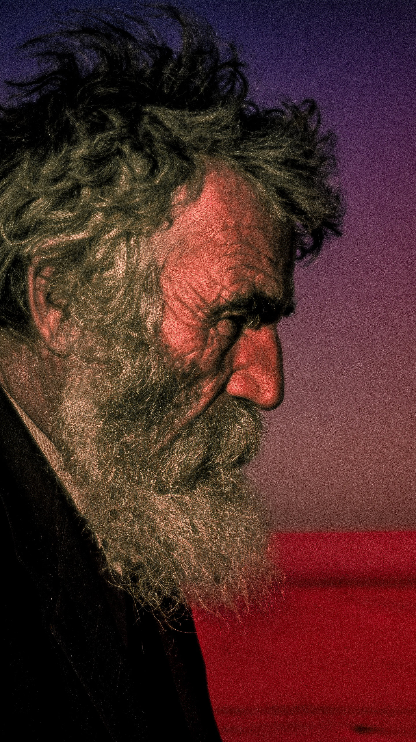 A close portrait of an older bearded man lit by blue and red light.