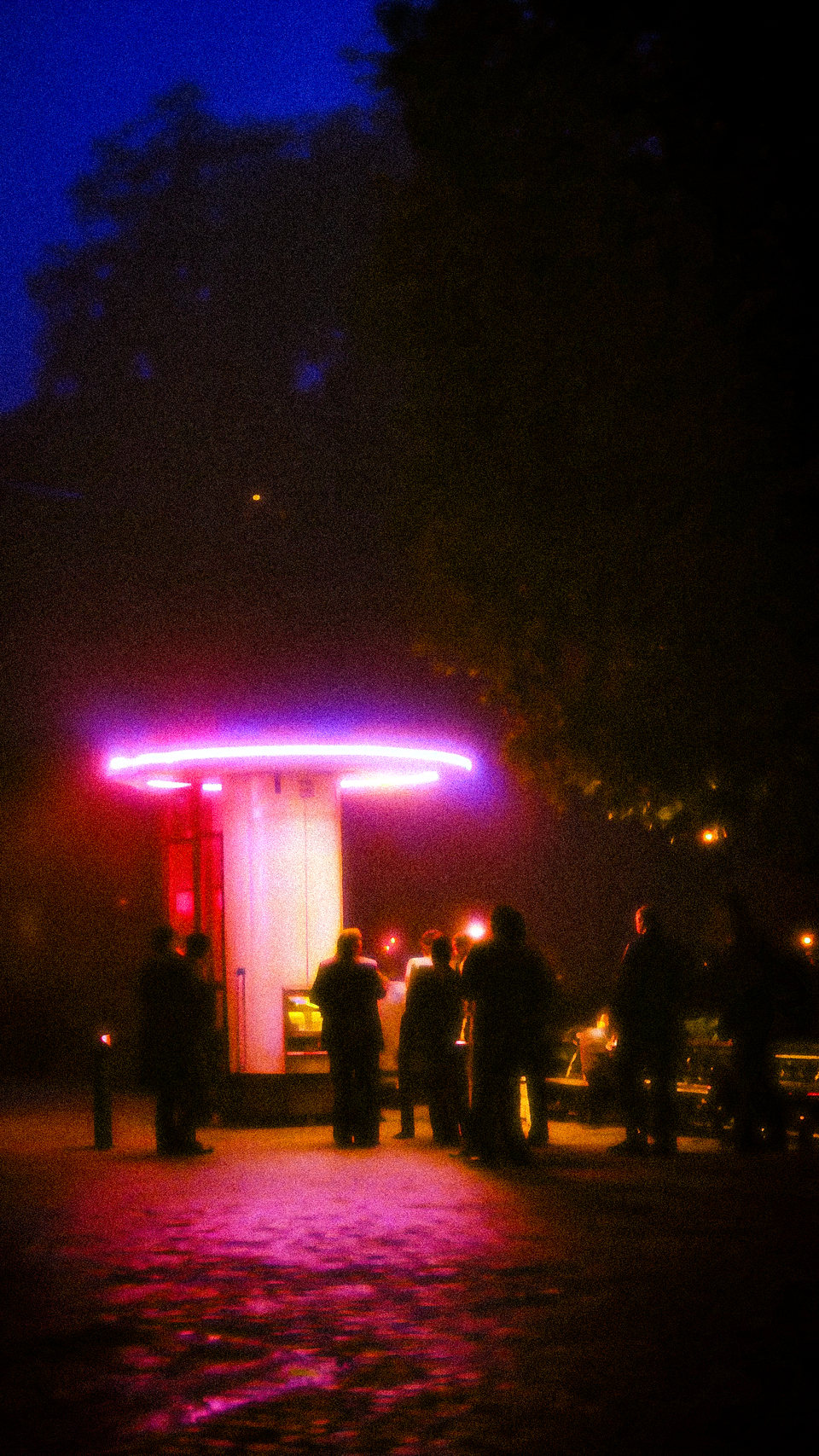 People gathered around a glowing pink kiosk in a dark park.